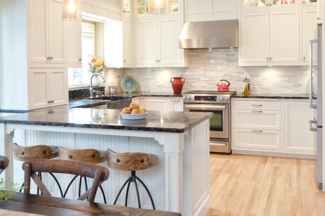 White kitchen with a dark stone island, carved wood counter stools, and a stainless range