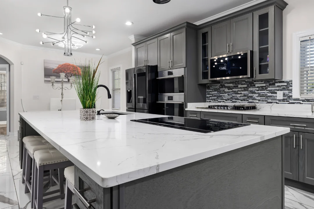 Gray kitchen cabinets with a white marble island and a flat black cooktop