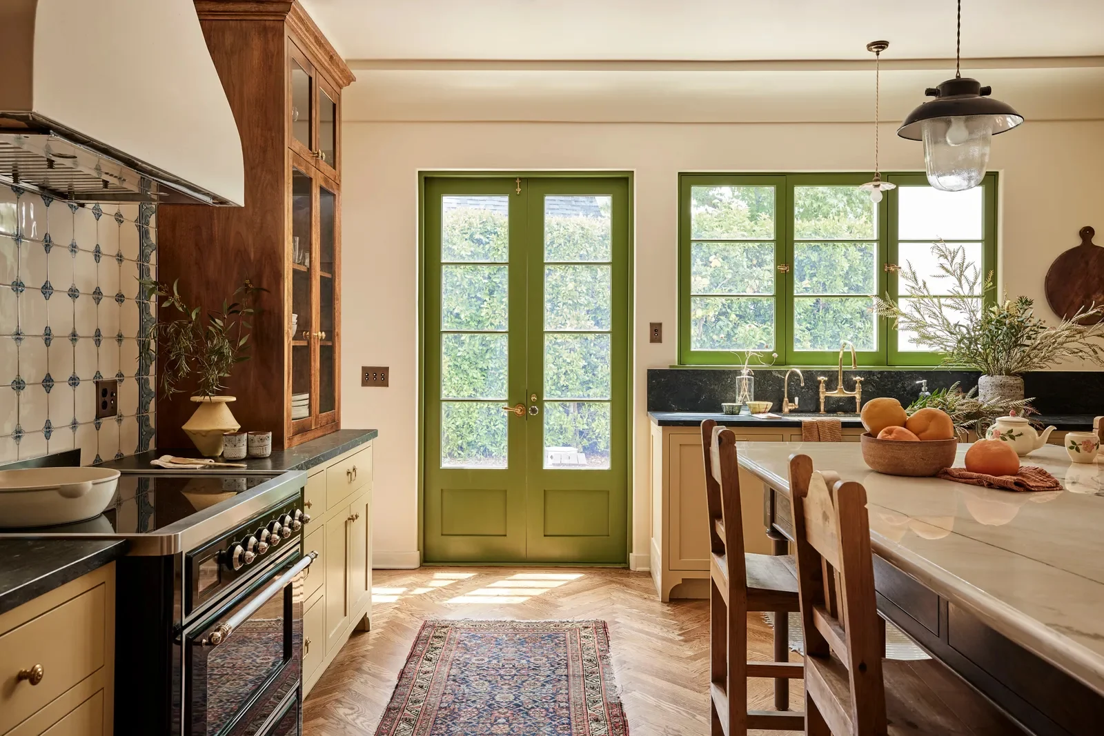 A Spanish Colonial Revival kitchen with green French doors, hand-painted tile behind the range, a plaster hood, and a herringbone wood floor